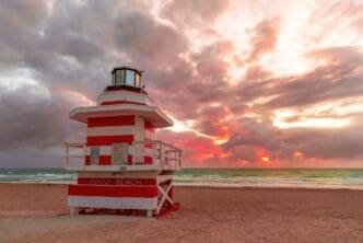A red and white striped lifeguard tower stands prominently on a sandy beach, facing the ocean where the sun sets with vibrant orange and red hues breaking through dramatic clouds. The sign on the tower reads "JETTY MIAMI BEACH".