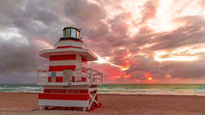 A red and white striped lifeguard tower stands prominently on a sandy beach, facing the ocean where the sun sets with vibrant orange and red hues breaking through dramatic clouds. The sign on the tower reads "JETTY MIAMI BEACH".