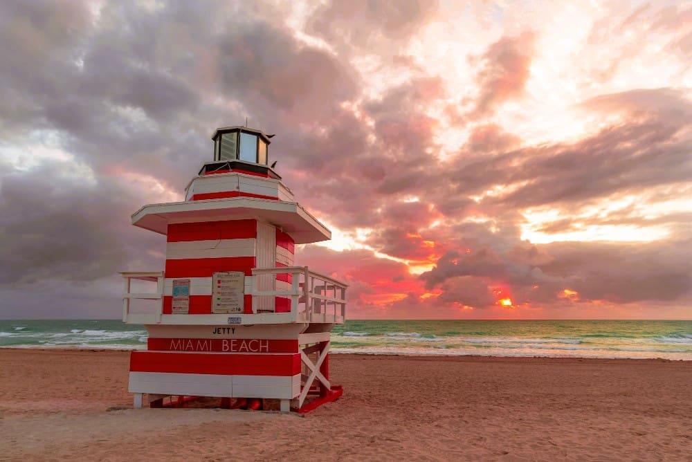 A red and white striped lifeguard tower stands prominently on a sandy beach, facing the ocean where the sun sets with vibrant orange and red hues breaking through dramatic clouds. The sign on the tower reads "JETTY MIAMI BEACH".