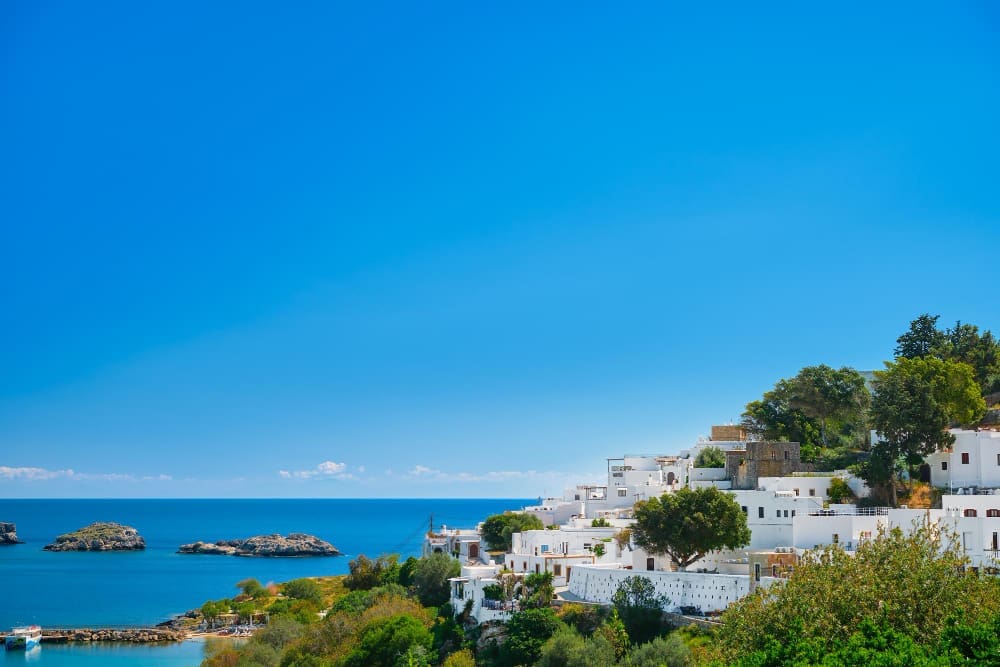 An elevated view of the ancient city of Lindos on the Greek island of Rhodes, showing its snow-white houses cascading down a hillside towards a deep blue bay with rocky islets in the Aegean Sea under a clear sky.