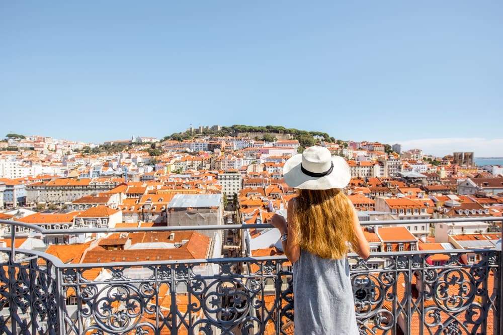 A young woman in a straw hat viewing the extensive cityscape of Lisbon, Portugal, with red-tiled roofs and a distant castle.