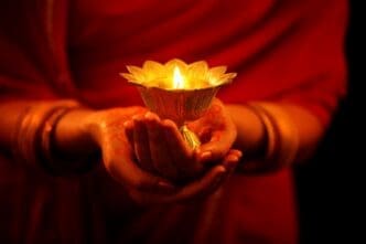 A woman's hands, adorned with bangles and mehndi, holding a glowing traditional lit diya (oil lamp).