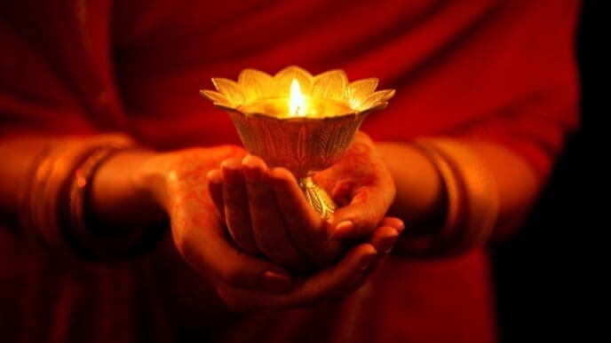 A woman's hands, adorned with bangles and mehndi, holding a glowing traditional lit diya (oil lamp).