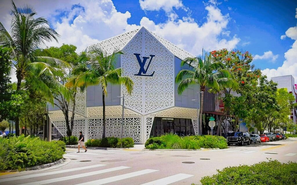 The Louis Vuitton building in the Miami Design District, featuring a white geometric facade with a large blue "LV" logo, surrounded by lush tropical landscaping and palm trees under a blue sky with fluffy clouds.