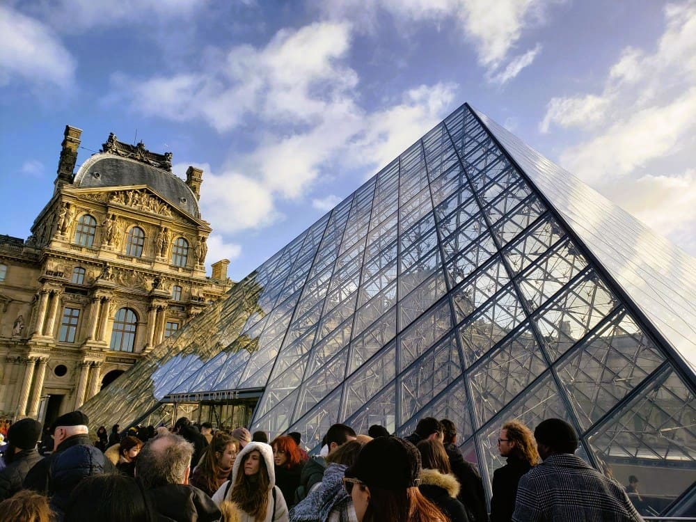 The modern glass Louvre Pyramid stands in front of the historic Louvre Palace in Paris, France, with a dense crowd of people gathered around its base under a cloudy sky.