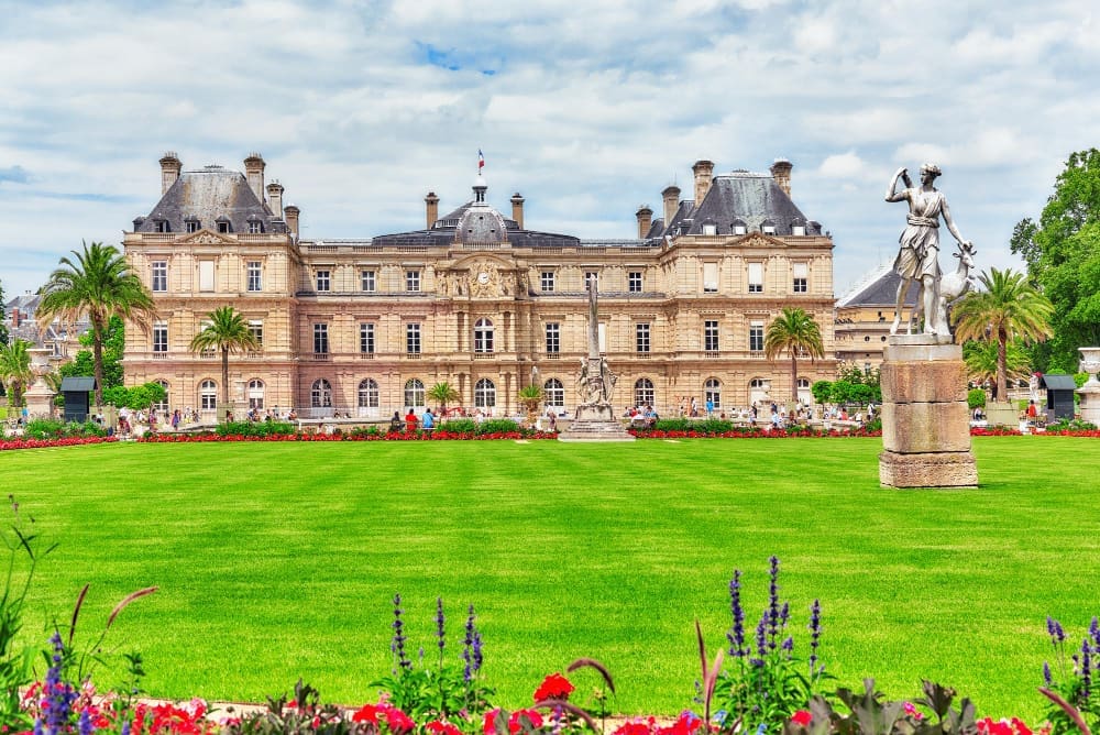 A wide view of the Luxembourg Palace in Paris, France, from across a vast green lawn with flowerbeds and a classical statue in the foreground, under a partly cloudy sky.