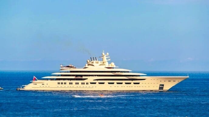 A massive beige mega yacht with a helicopter on its deck, moored in the sea on a sunny day.