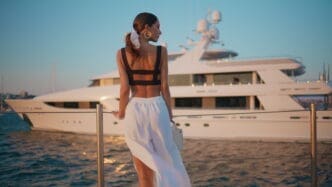 A woman in elegant resort wear standing by a railing, looking out at a luxurious superyacht on the water at sunset.