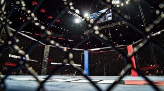 A low-angle view from inside an empty mixed martial arts (MMA) octagon, looking out through the chain-link fence towards bright arena lights and a jumbotron displaying fight information.