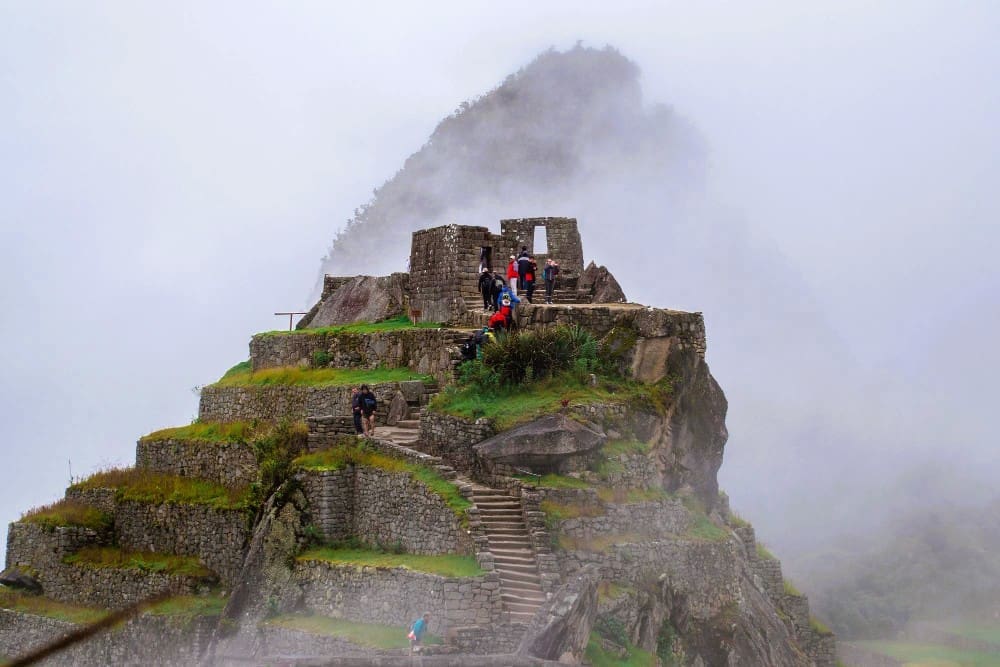 Ancient stone structures and terraced paths of Machu Picchu, partially shrouded in mist or low clouds, with people exploring the site.