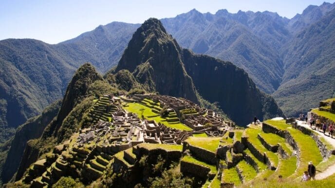 An elevated view of the ancient Inca city of Machu Picchu, with its terraces and structures, surrounded by dramatic green mountains under a clear sky.