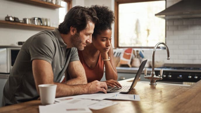 A focused couple looking at a laptop and documents on a kitchen counter, appearing to be managing household bills.