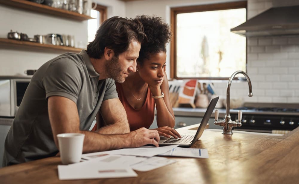 A focused couple looking at a laptop and documents on a kitchen counter, appearing to be managing household bills.