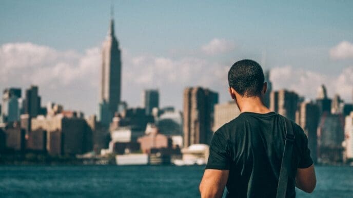 Rear view of a man standing by a body of water, looking at the distant Manhattan skyline under a sunny sky.