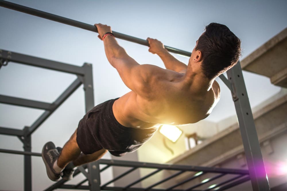 A shirtless man performing an intense pull-up or muscle-up exercise on a horizontal bar in an outdoor calisthenics park.