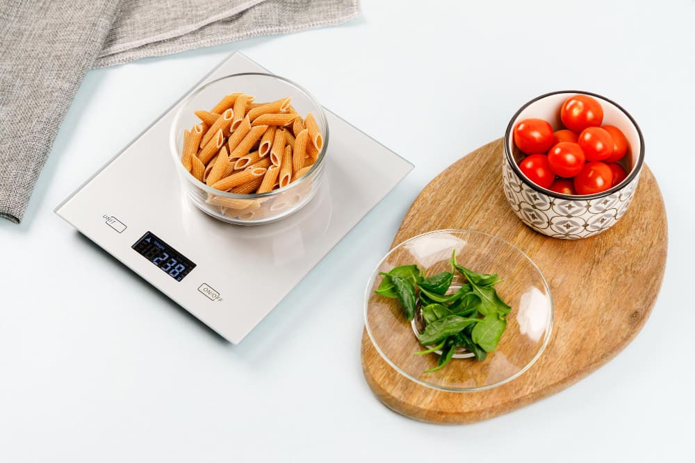 A glass bowl of dry penne pasta on a digital kitchen scale, next to cherry tomatoes and spinach, on a light surface.