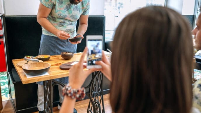 A man with a beard, wearing a green t-shirt and apron, prepares food at a small wooden table. In the foreground, a person, seen from behind, takes a picture of the scene with their smartphone, illustrating a food blogger.