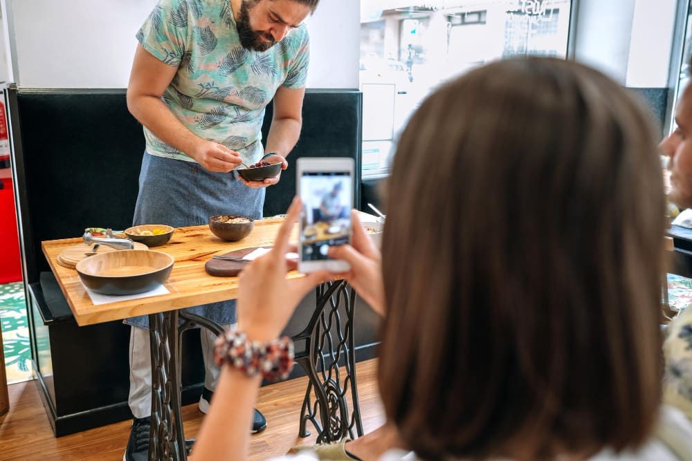 A man with a beard, wearing a green t-shirt and apron, prepares food at a small wooden table. In the foreground, a person, seen from behind, takes a picture of the scene with their smartphone, illustrating a food blogger.