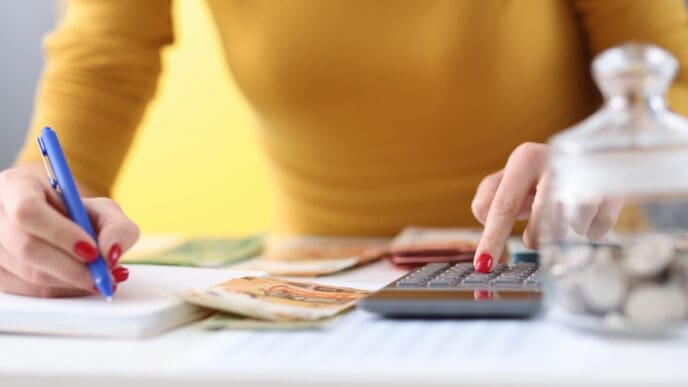A close-up of a person's hands using a calculator and writing in a notebook, with banknotes and a glass jar of coins on the desk, symbolizing budgeting and money management.