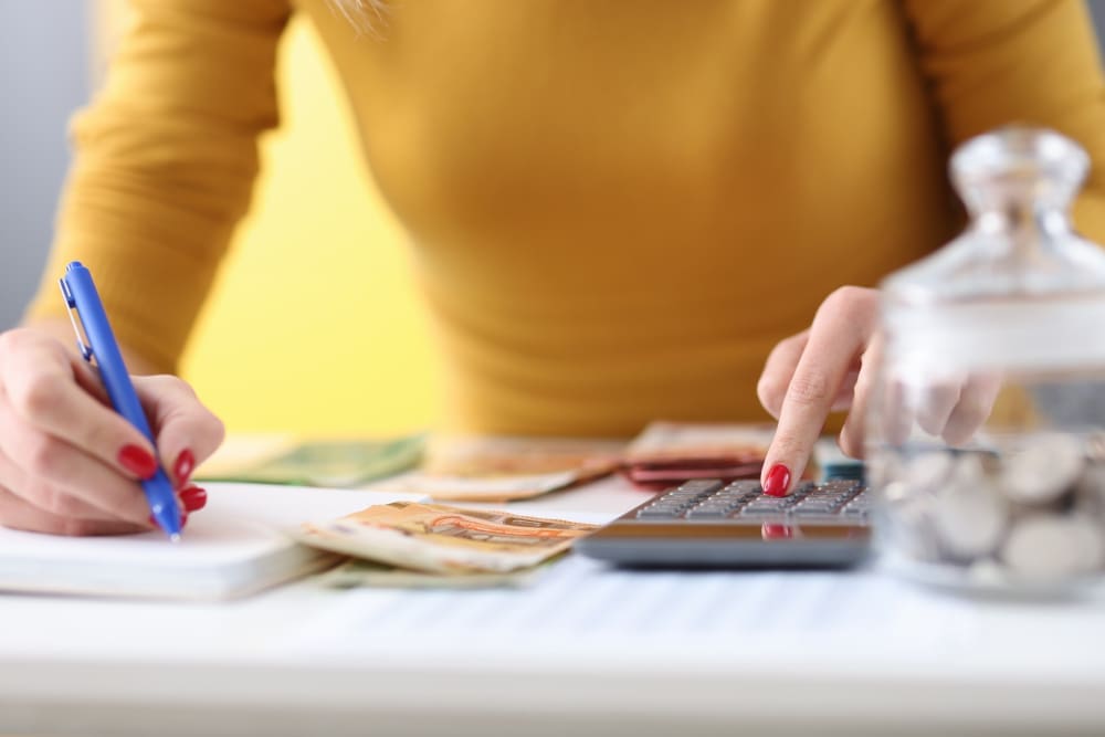 A close-up of a person's hands using a calculator and writing in a notebook, with banknotes and a glass jar of coins on the desk, symbolizing budgeting and money management.