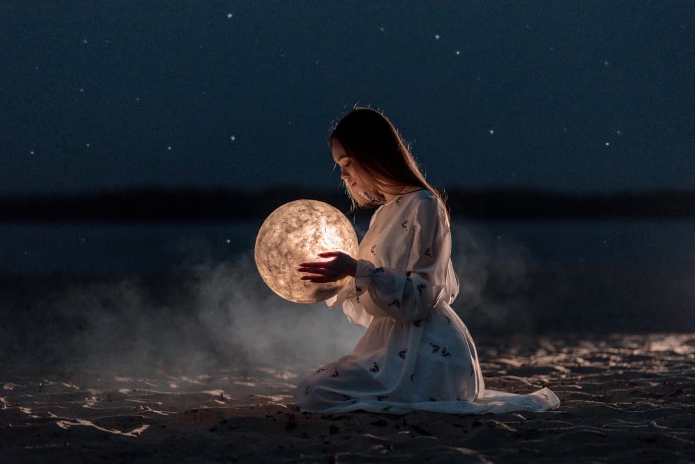 A young woman in a white dress kneels on a dark sandy surface at night, gently holding a glowing, textured sphere resembling the moon, with faint stars in the dark sky and a blurred body of water in the background.