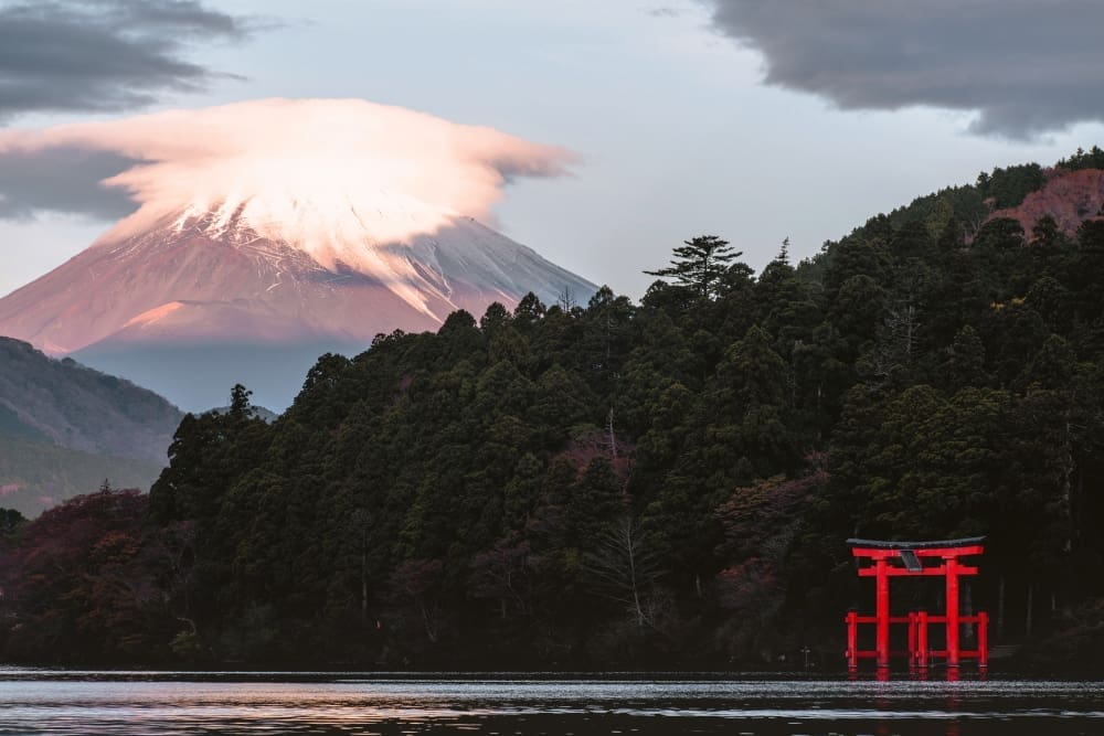 Mount Fuji, topped with a cloud, rises majestically behind Lake Ashi, where a red torii gate stands partially submerged in the foreground, surrounded by a forested shoreline.