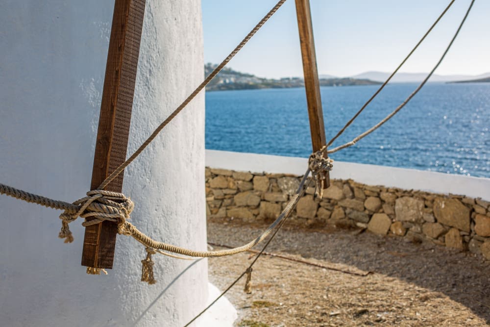 A selective focus shot of the lower part of a whitewashed windmill tower in Mykonos, Greece, showing wooden beams and ropes in the foreground, with the deep blue Aegean Sea and distant land in the blurred background.