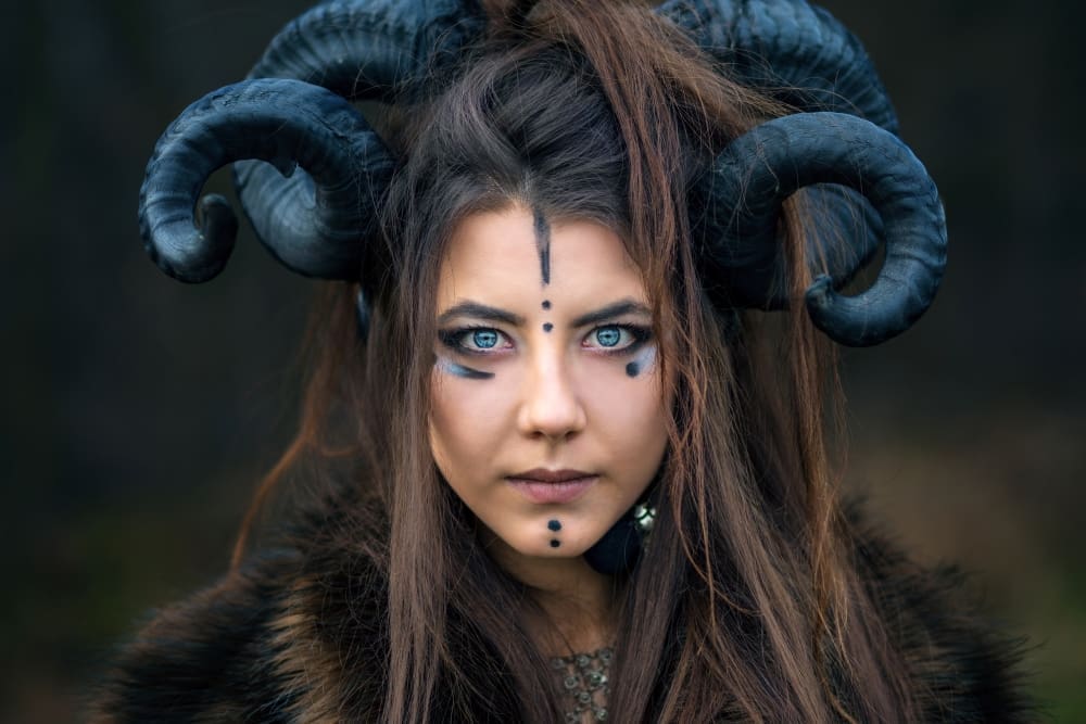Close-up portrait of a serious woman with dramatic eye makeup, painted face markings, and large black ram horns.
