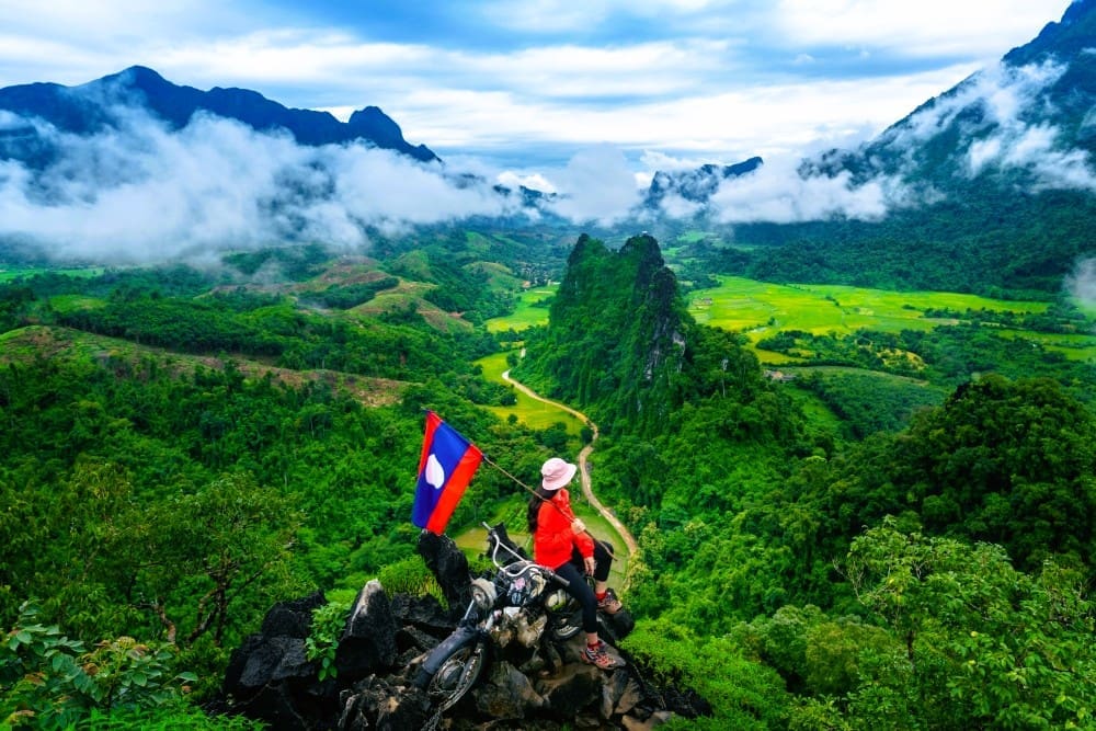 A tourist holding a Lao flag while sitting on a motorbike at Nam Xay Viewpoint, overlooking lush green mountains and valleys with mist, in Vang Vieng, Laos.