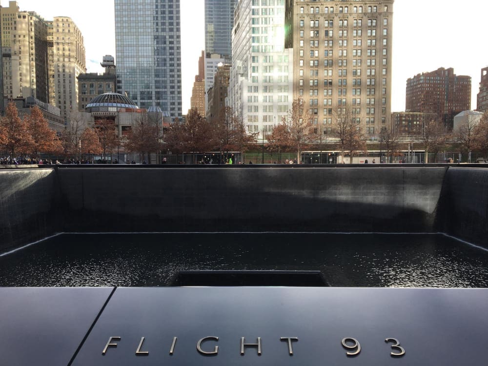 The North Pool of the National September 11 Memorial, with the words "FLIGHT 93" inscribed on the bronze parapet in the foreground, and modern skyscrapers in the background under a bright sky.