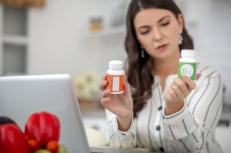 A woman with dark hair holds up two white supplement bottles, one with an orange label and another labeled "B12," and looks at them with a contemplative expression, with a laptop in the blurred foreground, symbolizing health decisions.