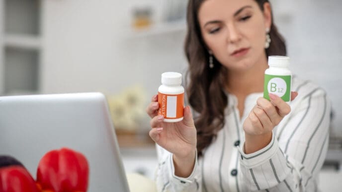 A woman with dark hair holds up two white supplement bottles, one with an orange label and another labeled "B12," and looks at them with a contemplative expression, with a laptop in the blurred foreground, symbolizing health decisions.