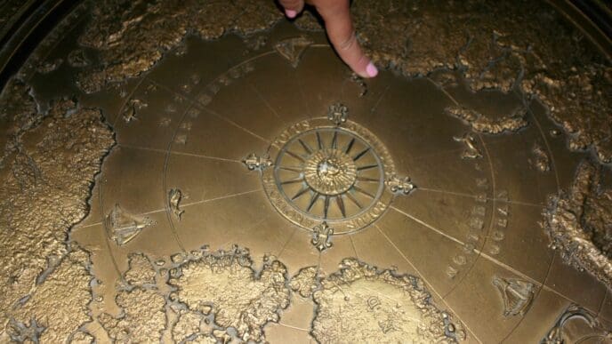 A hand with a pink fingernail points to a detailed brass relief map featuring continents, oceans, and a central compass rose, appearing to be part of a large, circular, antique-style globe or monument.