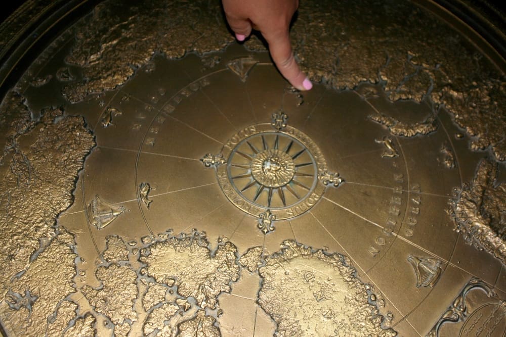 A hand with a pink fingernail points to a detailed brass relief map featuring continents, oceans, and a central compass rose, appearing to be part of a large, circular, antique-style globe or monument.