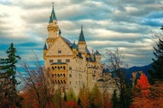 The majestic Neuschwanstein Castle, a white stone fairytale palace with blue roofs and multiple turrets, stands tall amidst colorful autumn trees and a dramatic cloudy sky.