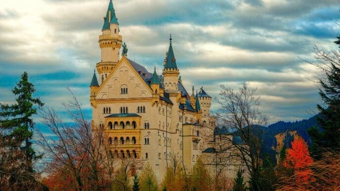 The majestic Neuschwanstein Castle, a white stone fairytale palace with blue roofs and multiple turrets, stands tall amidst colorful autumn trees and a dramatic cloudy sky.