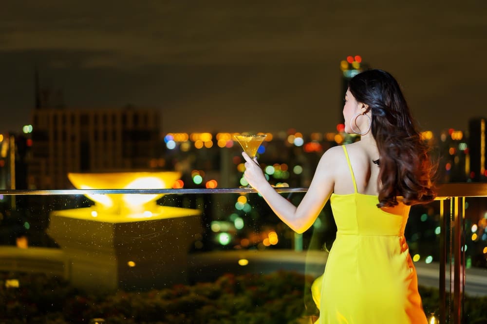 A stylish woman in a bright yellow dress stands on a rooftop bar at night, holding a martini glass, and looks out at the illuminated city skyline.