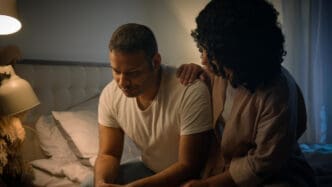 A woman gently comforting a man who appears distressed, with his head down, in a dimly lit bedroom.
