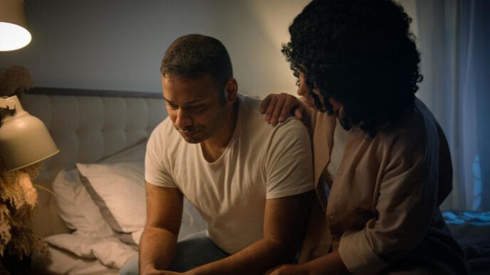 A woman gently comforting a man who appears distressed, with his head down, in a dimly lit bedroom.