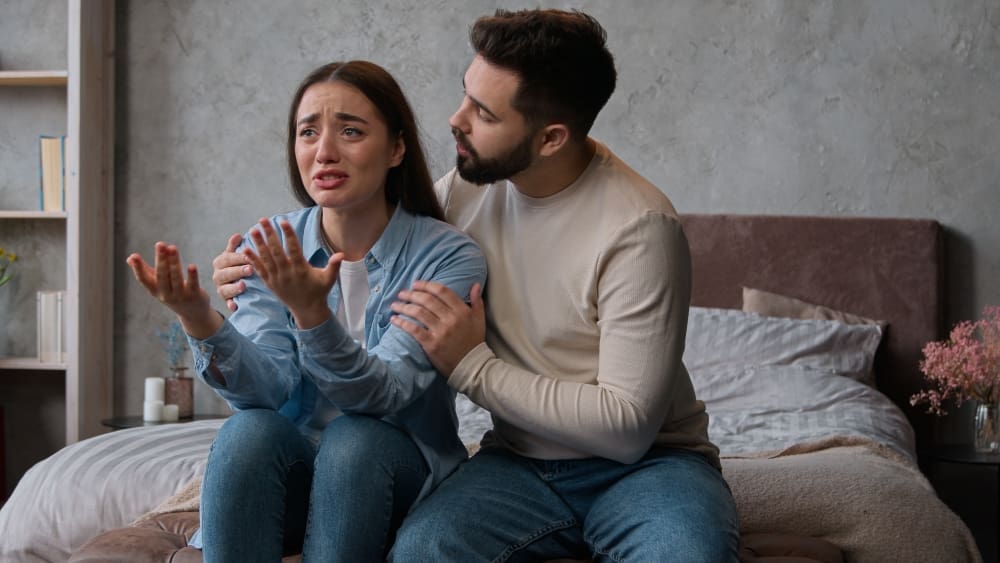 A man comforting his distressed wife, who is crying and gesturing, possibly after a conflict, in a bedroom setting.