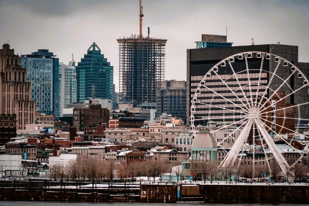 The Montreal skyline, featuring a large white Ferris wheel, modern skyscrapers, and older buildings, set against a grey sky, with a body of water and foreground showing hints of winter.