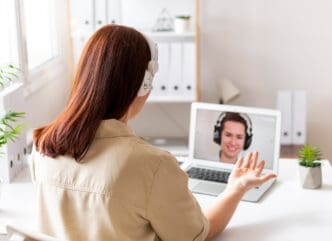 A woman seen from behind, wearing white headphones, engaged in a video call on her laptop with a smiling therapist also wearing headphones, illustrating an online therapy session.