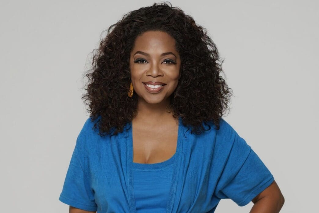 A smiling Oprah Winfrey, with her signature curly dark hair, wearing a vibrant blue top and cardigan, looks directly at the camera.