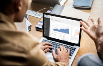 An over-the-shoulder view of a person typing on a laptop, which displays a business chart and visual data, with another person's gesturing hands visible to the right, in a collaborative work setting.