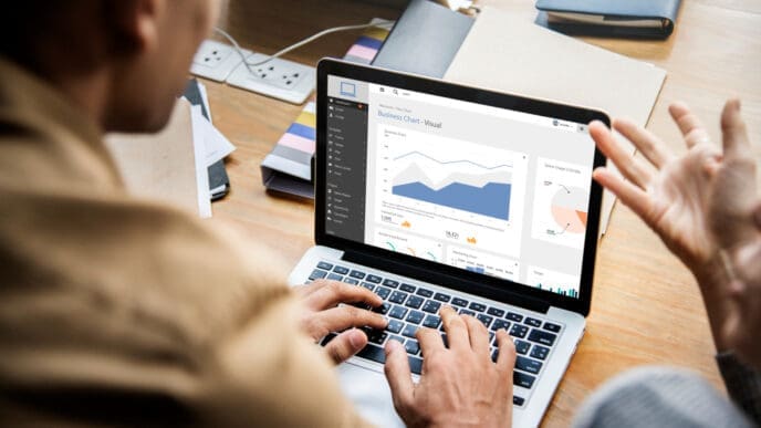 An over-the-shoulder view of a person typing on a laptop, which displays a business chart and visual data, with another person's gesturing hands visible to the right, in a collaborative work setting.