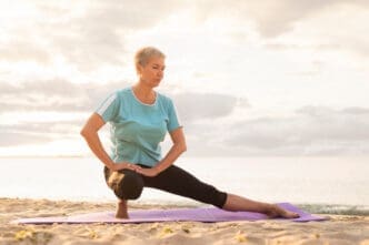 An older woman with short gray hair performs a side lunge stretch on a purple yoga mat on a sandy beach, with the ocean and a hazy sky in the background at sunrise or sunset.