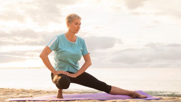 An older woman with short gray hair performs a side lunge stretch on a purple yoga mat on a sandy beach, with the ocean and a hazy sky in the background at sunrise or sunset.