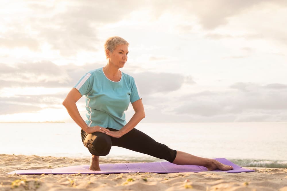 An older woman with short gray hair performs a side lunge stretch on a purple yoga mat on a sandy beach, with the ocean and a hazy sky in the background at sunrise or sunset.