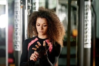 A woman with curly, dark hair, wearing a black and pink athletic top, looks down intently as she adjusts a fitness glove on her hand in a gym setting.