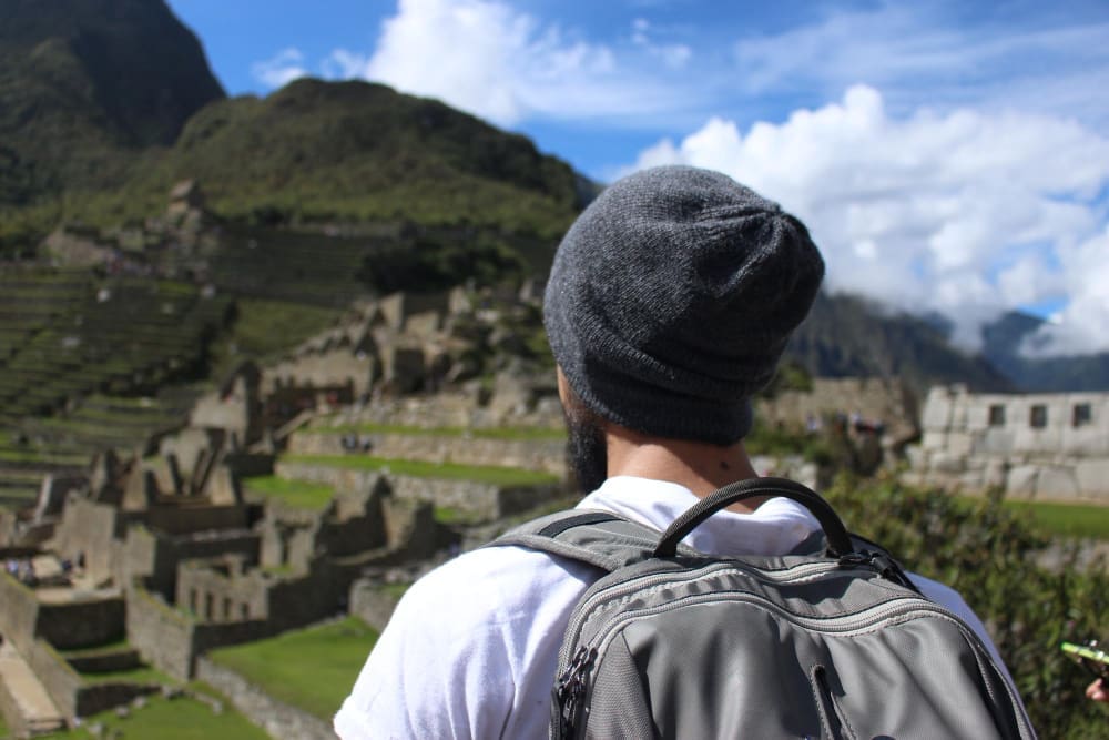 A man with a grey beanie and backpack stands with his back to the camera, overlooking the ancient Inca city of Machu Picchu with its terraced ruins and mountains under a blue, cloudy sky.